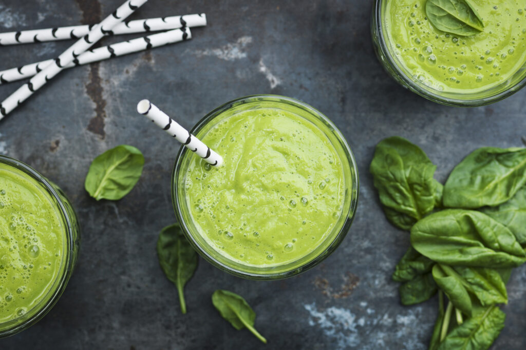 Auf einem Tisch steht ein Smoothie, daneben liegen Spinatblätter und Strohhalme.