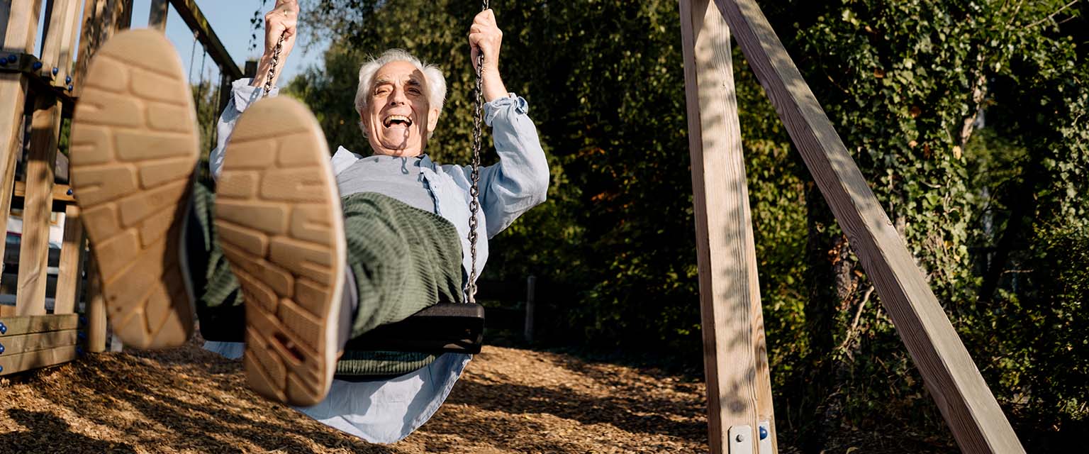 Ein alter Mann schaukelt auf dem Spielplatz und streckt lachend die Zunge heraus.