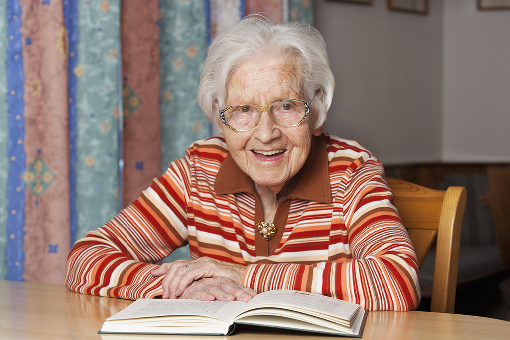 Eine alte Frau sitzt am Tisch, vor ihr liegt ein aufgeschlagenes Buch. Sie lächelt.