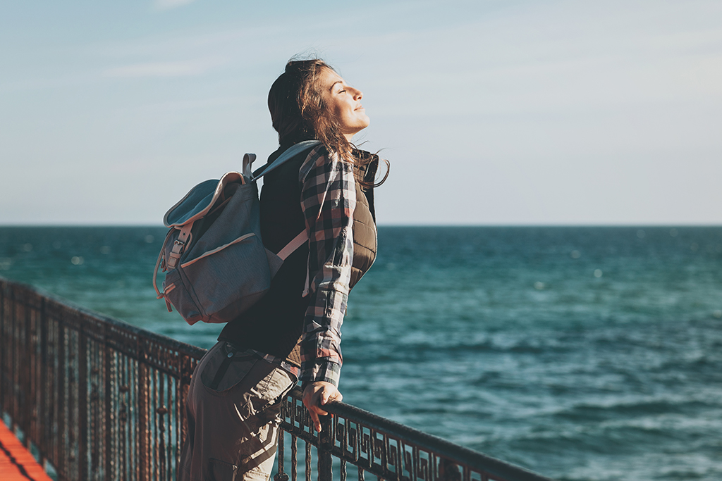 An einem Steg am Meer lehnt eine junge Frau am Zaun und hält das Gesicht in den Sonnenschein. Sie trägt einen Rucksack.