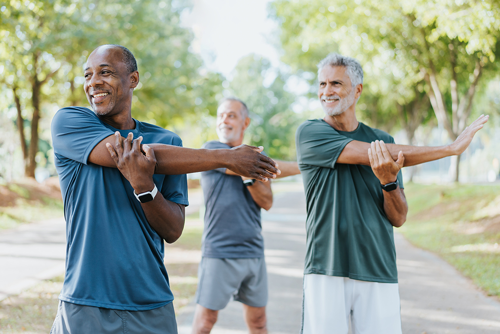 Drei ältere Männer ziehen mit der linken Hand dem rechten Oberarm nach vorn, um die Schulter zu dehnen. Sie lächeln.