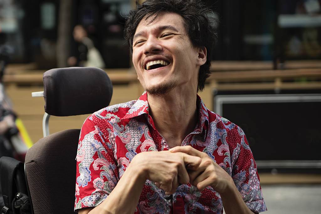 Ein im Rollstuhl sitzender junger Mann lächelt. Seine Hände sind verformt, die Finger gekrümmt.