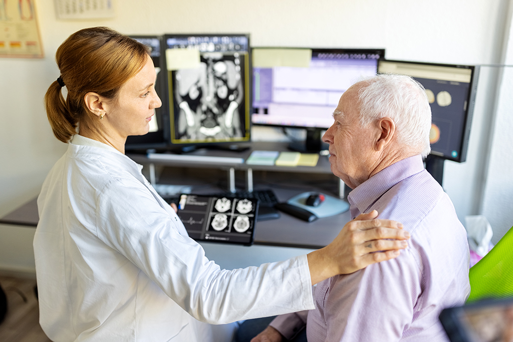 Eine Ärztin unterhält sich mit einem älteren Mann. Sie hat ihre rechte Hand auf seine Schulter gelegt. Beide stehen vor einem Bildschirm, der Röntgenbefunde zeigt.