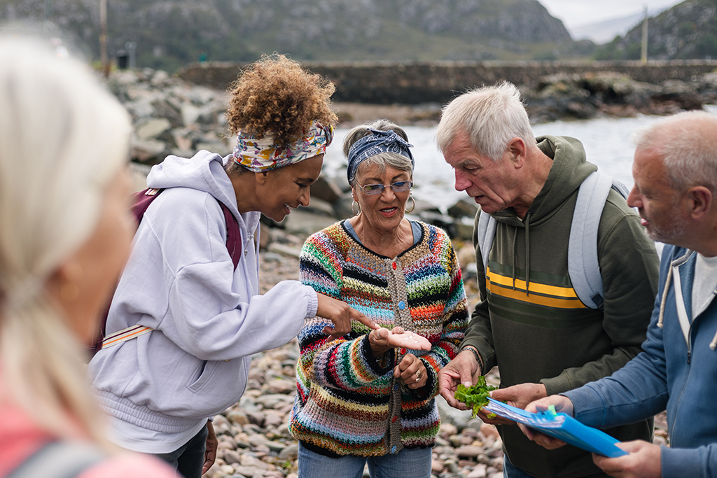 Zwei Frauen und zwei Männer schauen auf ein Fundstück, welches auf der Handfläche einer der Frauen liegt. Im Hintergrund ist ein See und Gebirge zu sehen.