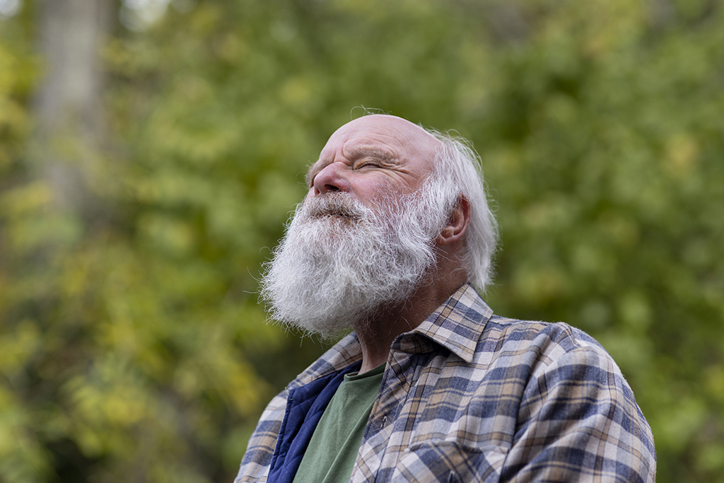 Ein alter Mann mit Bart steht mit geschlossenen Augen im Grünen. Er hält die Nase in die Luft.