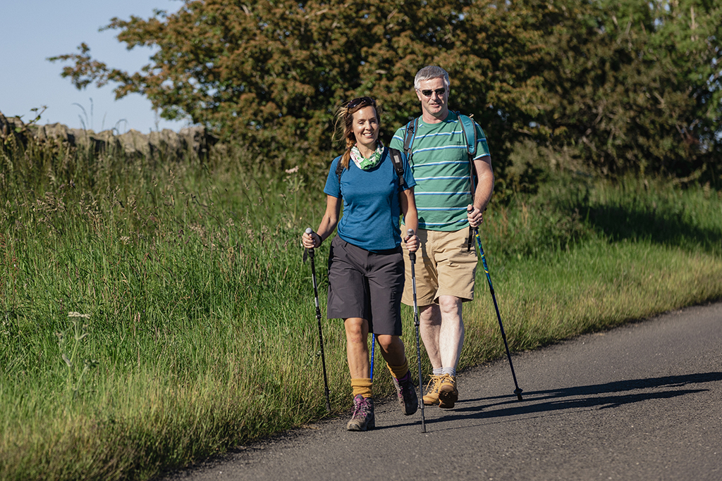 Eine Frau und ein Mann walken auf einem Feldweg. Sie verwenden Trecking-Stöcke.
