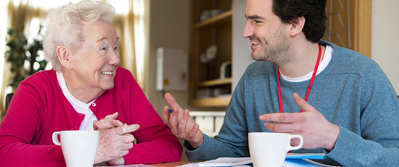 Eine alte Frau und ein junger Mann sitzen an einem Tisch und unterhalten sich. Beide lächeln.