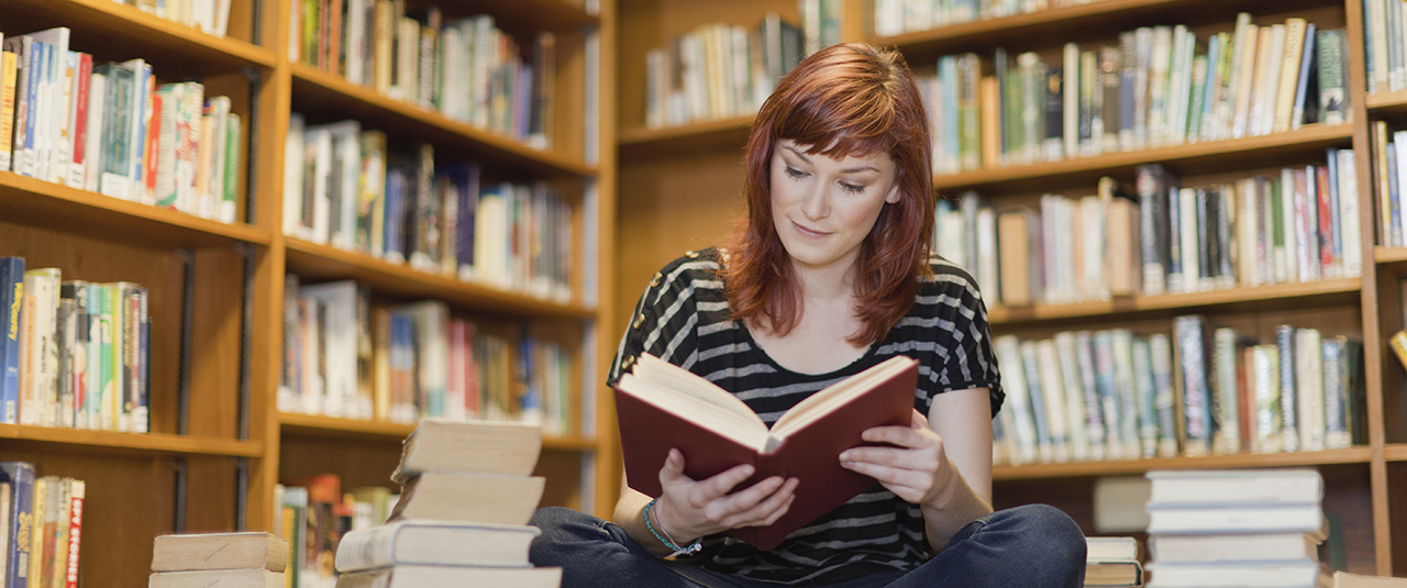 Eine junge Frau sitzt im Schneidersitz am Boden und liest in einem Buch. Neben ihr sind verschiedene Bücher gestapelt. Im Hintergrund stehen Regale voller Bücher.