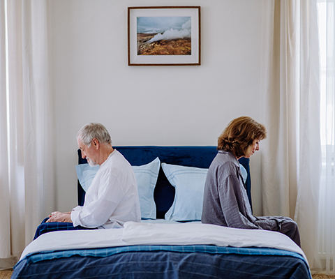 Eine älteres Paar sitzt Rücken an Rücken auf der Bettkante.