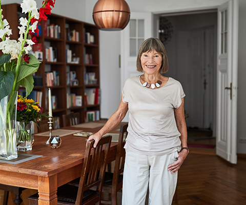 Eine Seniorin steht stolz und glücklich an ihrem Esstisch.